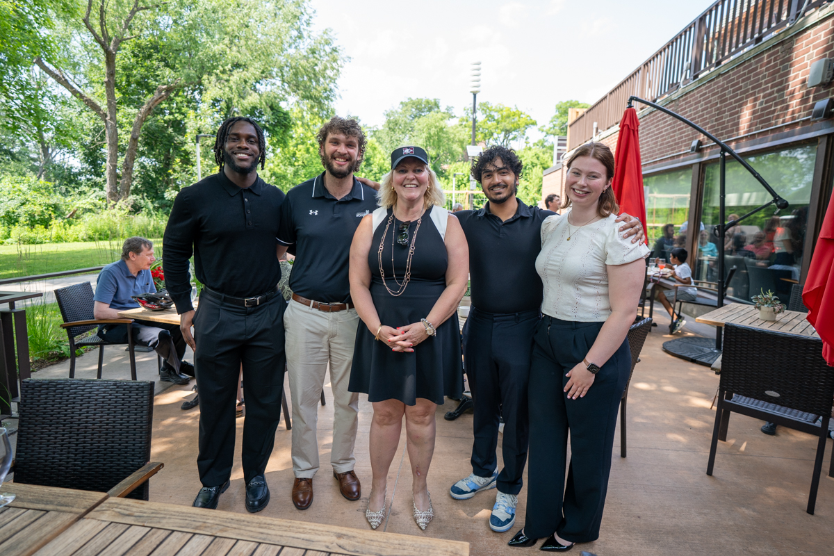 Four students and Susan Tighe stand together, smiling for a photo on the patio of Alumni Memorial Hall.