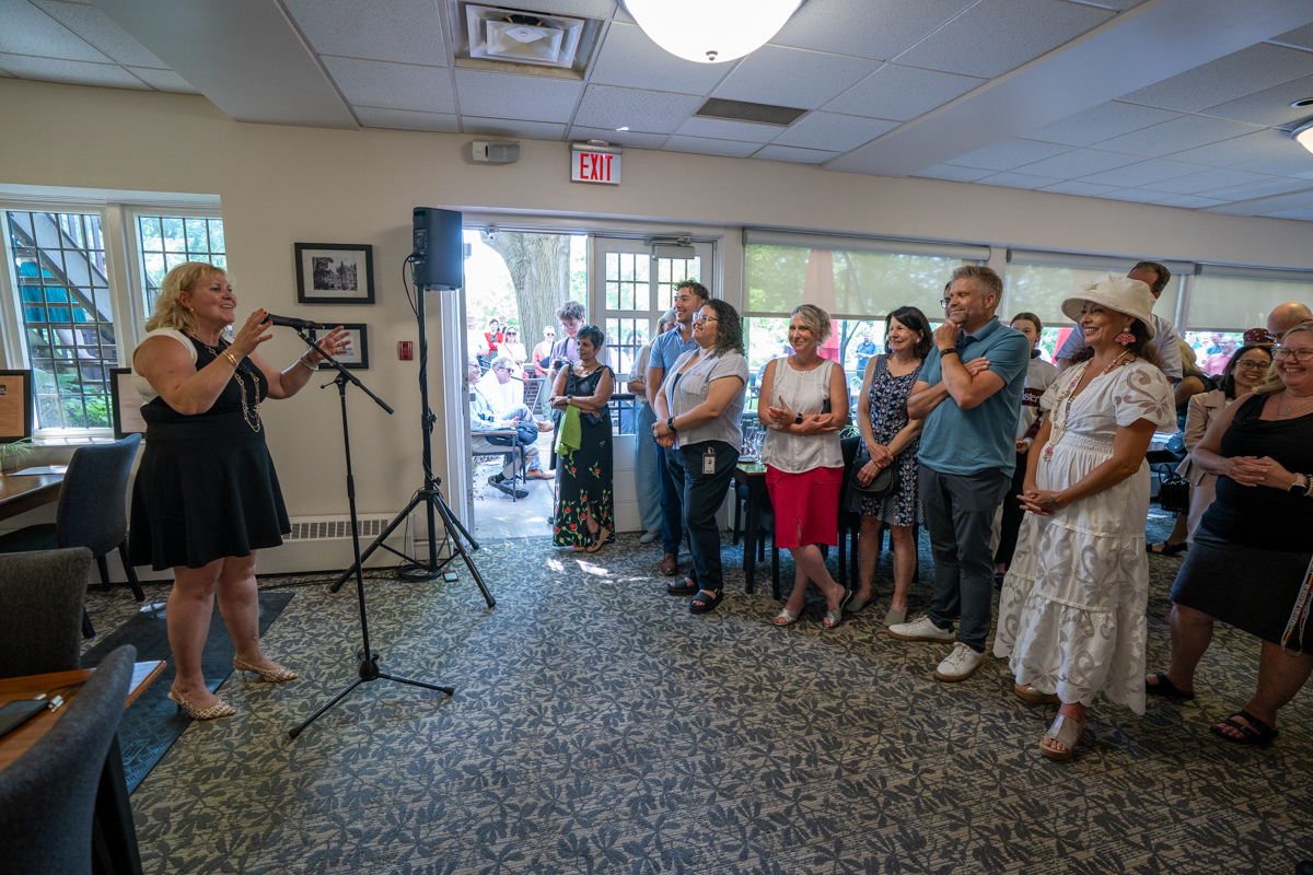Susan Tighe speaks into a microphone to a room full of people with doors that lead to a patio in the background.
