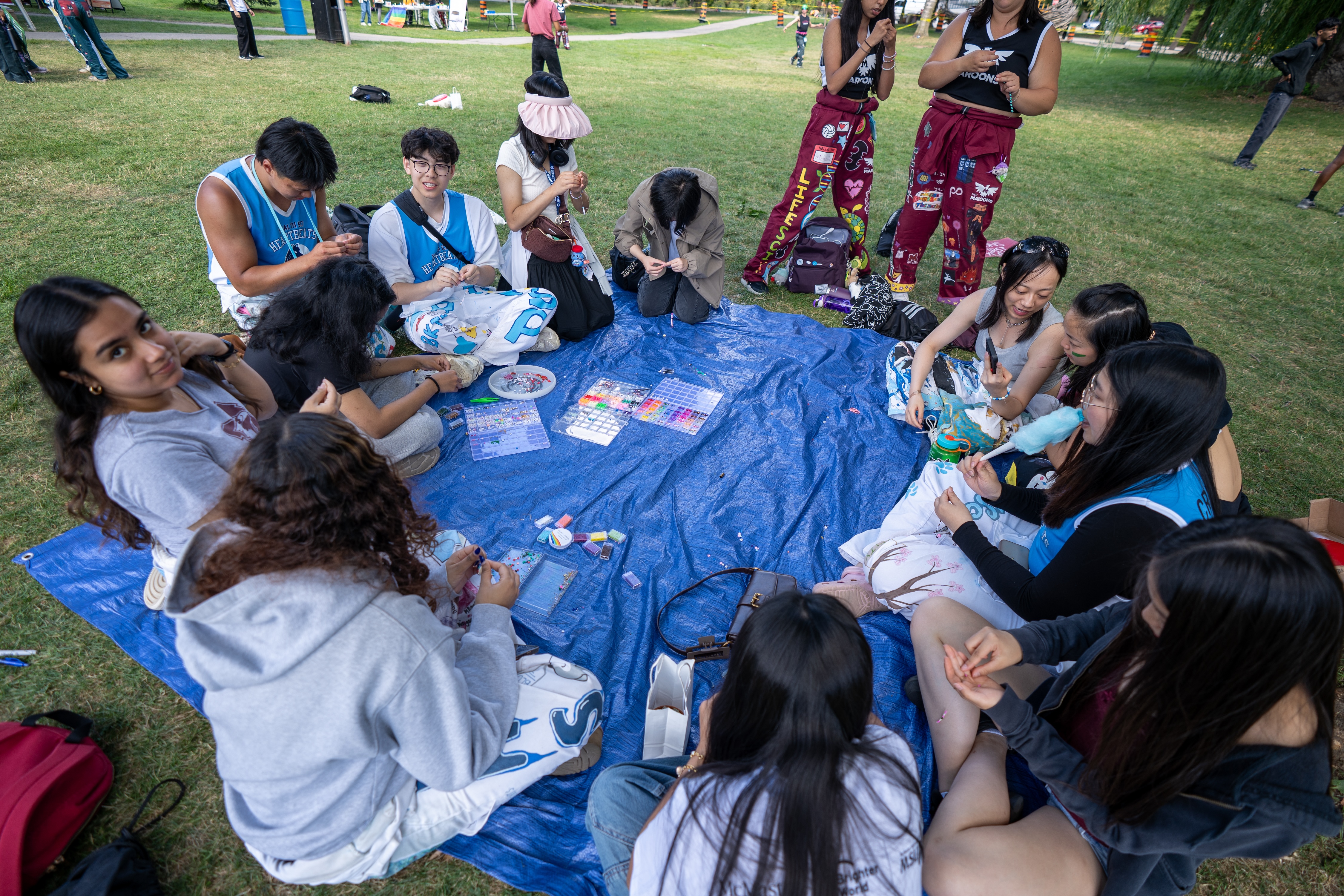 A group of students and welcome week reps sit around a big blue tarp laid out on a field, doing art and crafts