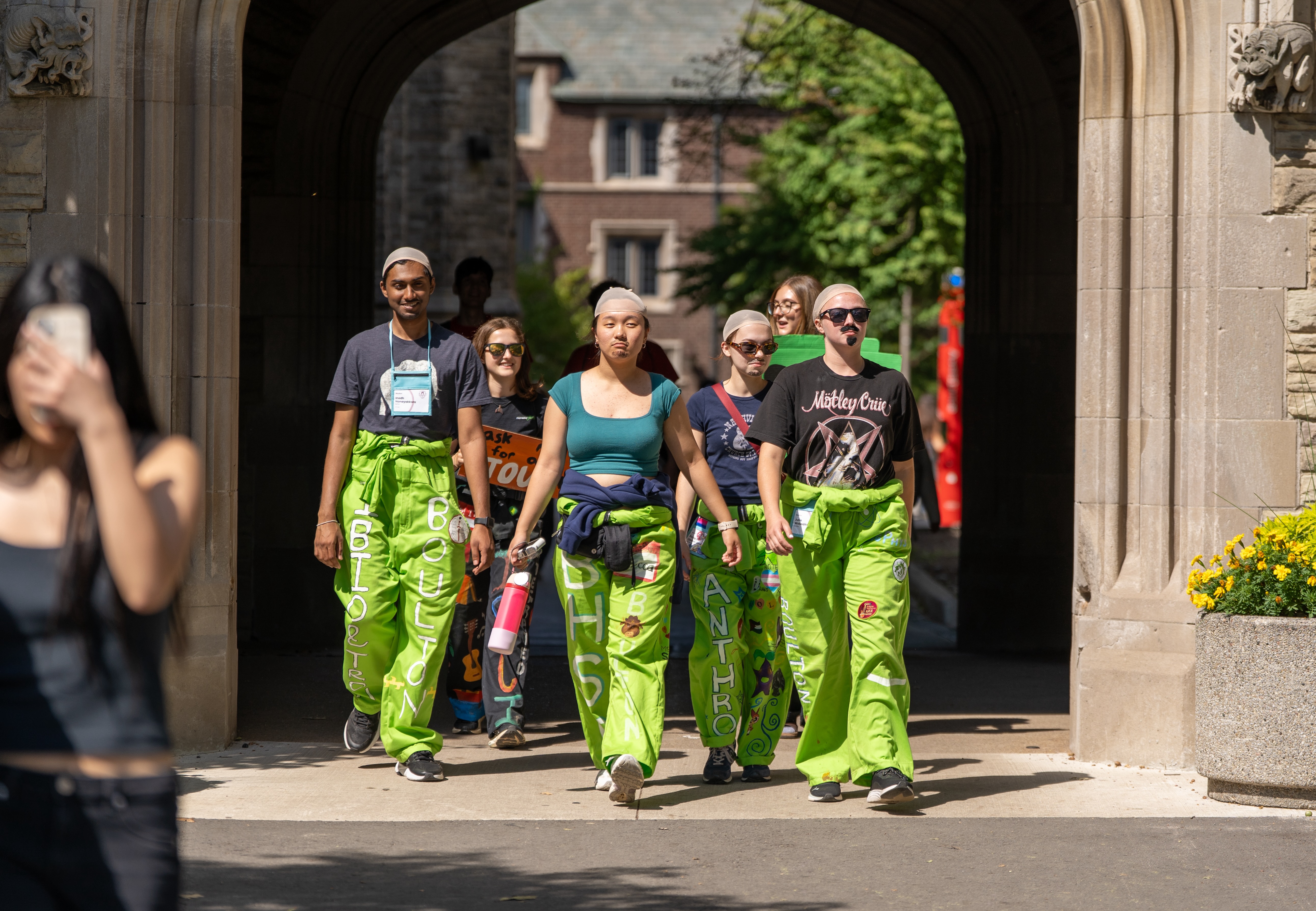 A group of welcome week reps in light green jumpsuits, some with fake facial hair, some with wig caps, walk out from under the University Hall archway.