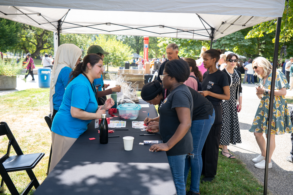 A table is set up under a shade tent, with several people gathered around it.