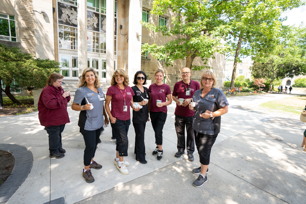 A group of people in maroon, grey and black uniforms stand outside, in front of a large building, holding ice creams. 