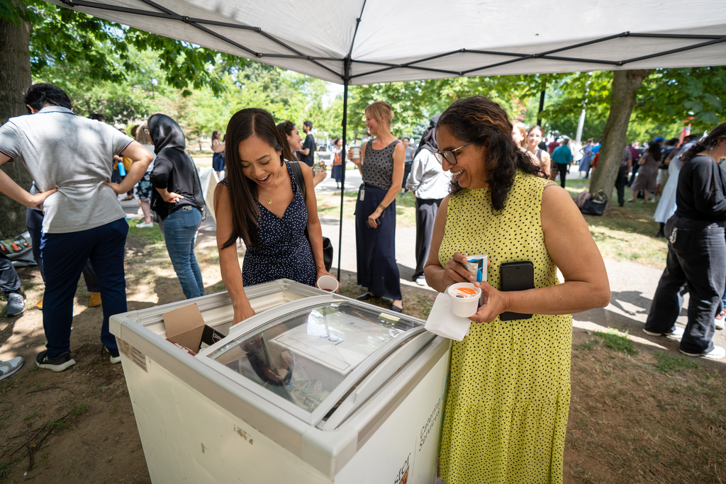 Standing outdoors under a canopy, two women lean over a freezer, holding ice cream cups.