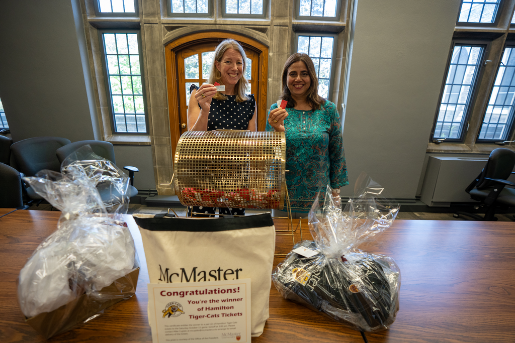 Two women stand at a wooden table, holding up raffle tickets. A ticket spinner and various prizes are on the table in front of them.