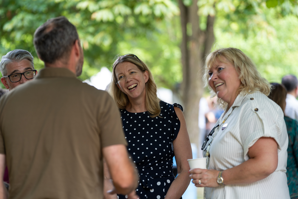 A group of people stand outside. Two women's faces are visible, laughing and tilted slightly. 