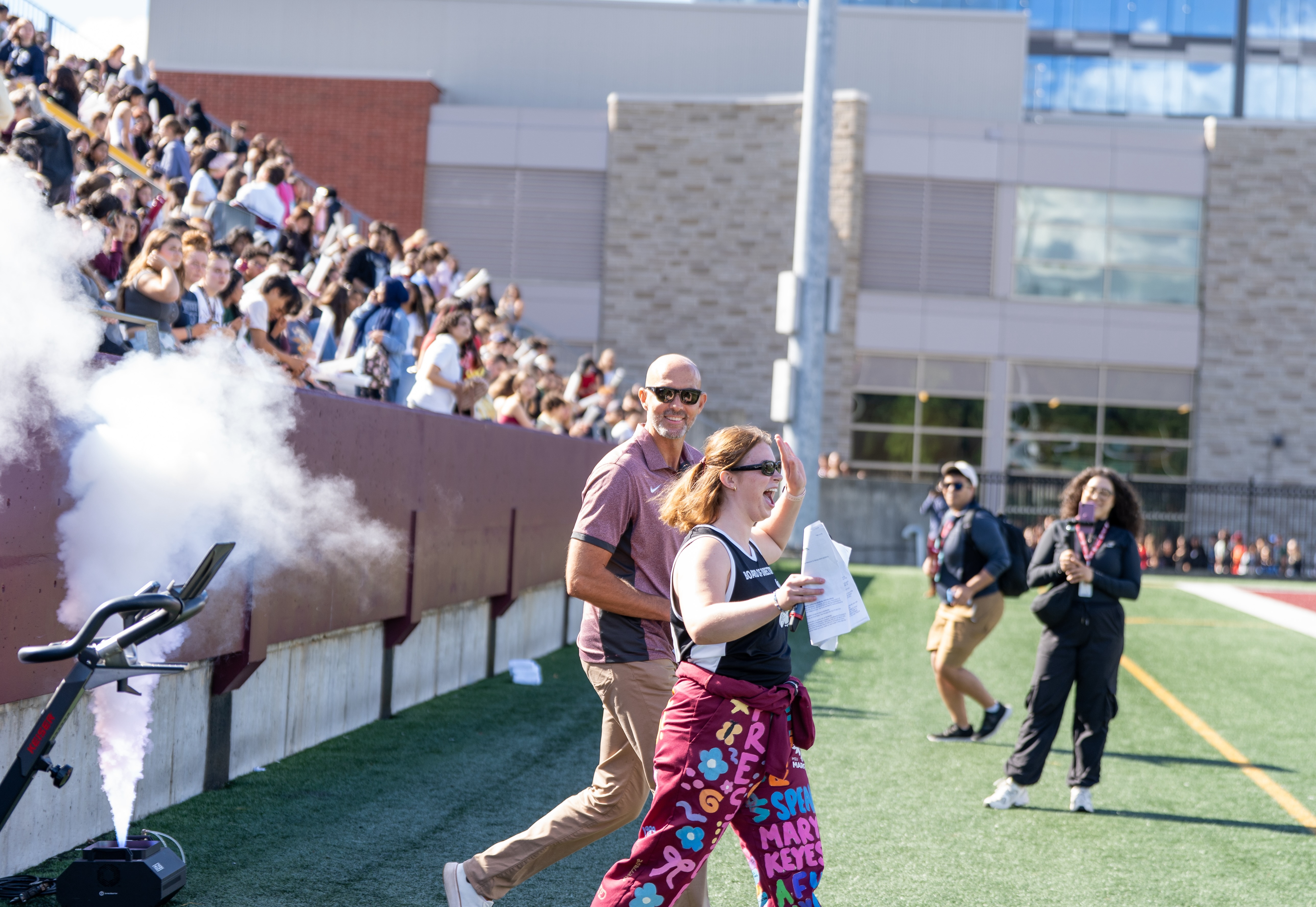 Smiling Sean Van Koughnett and Piper Plavins run out on to the field with smoke from a smoke machine behind them. The crowd in the stands is cheering.