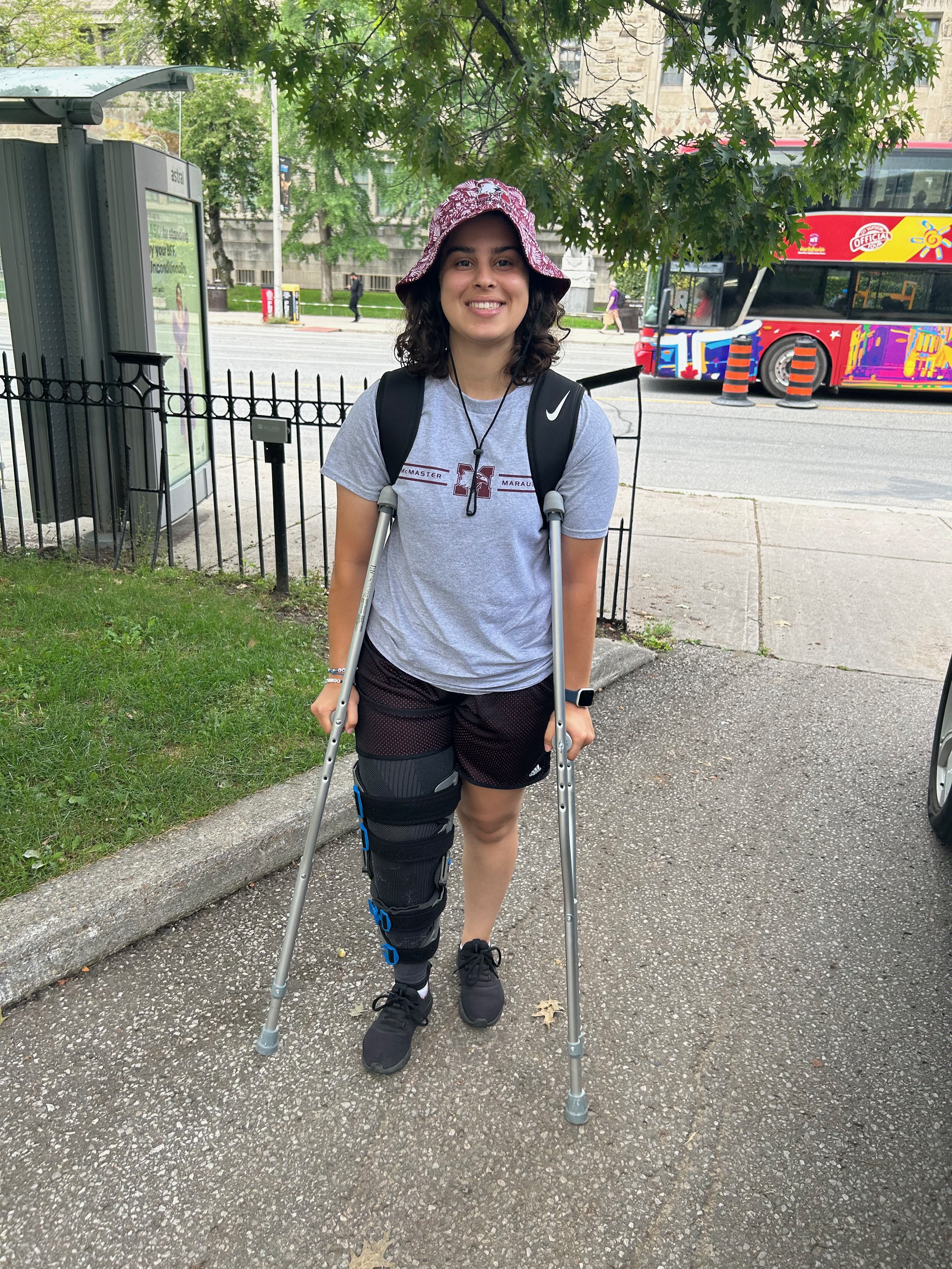 A woman stands on a sidewalk, using crutches, with a brace on her right knee and leg. 