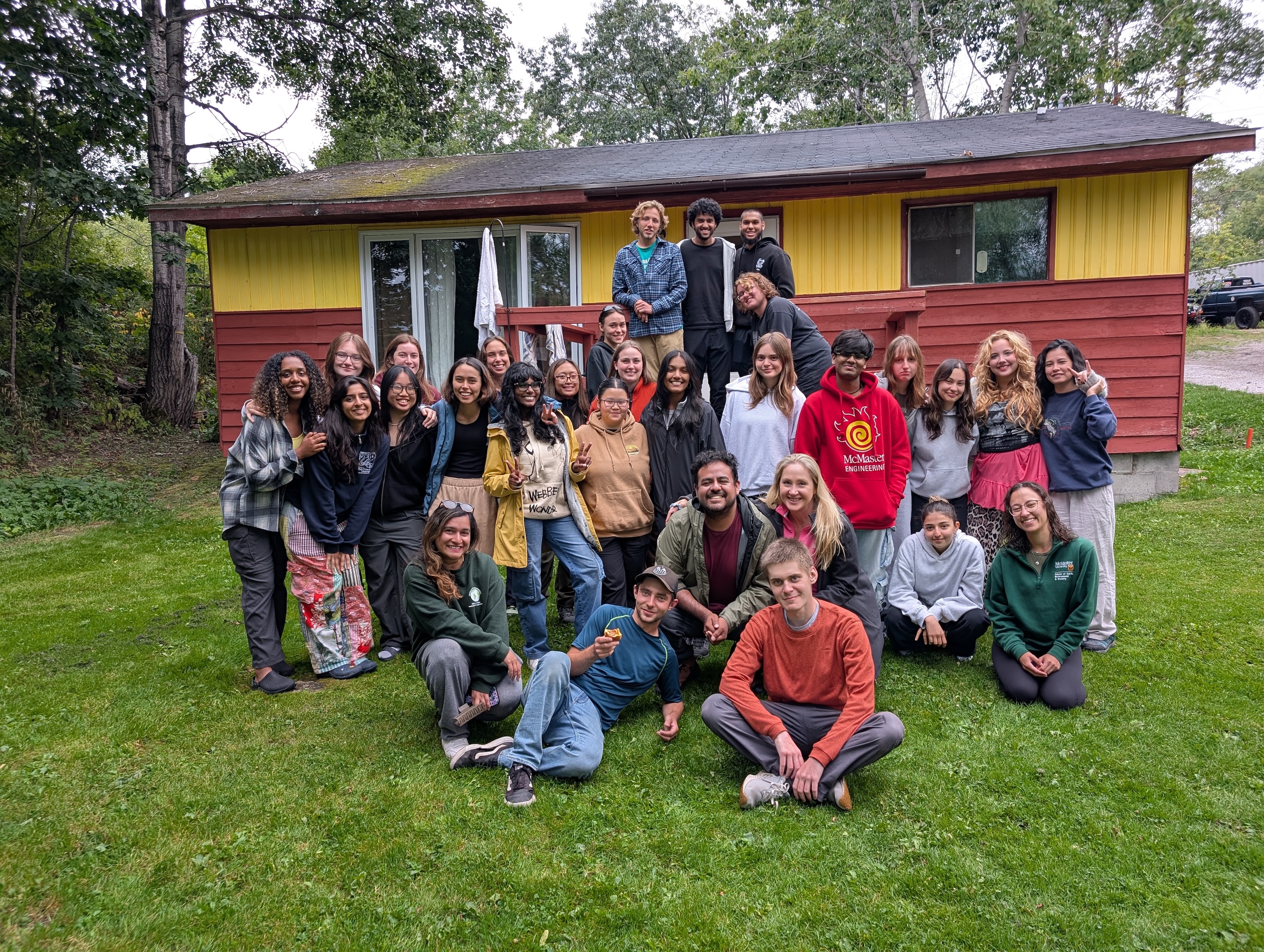 A group shot of many people, in jeans and hoodies, sitting and standing on the lawn outside of a cabin.