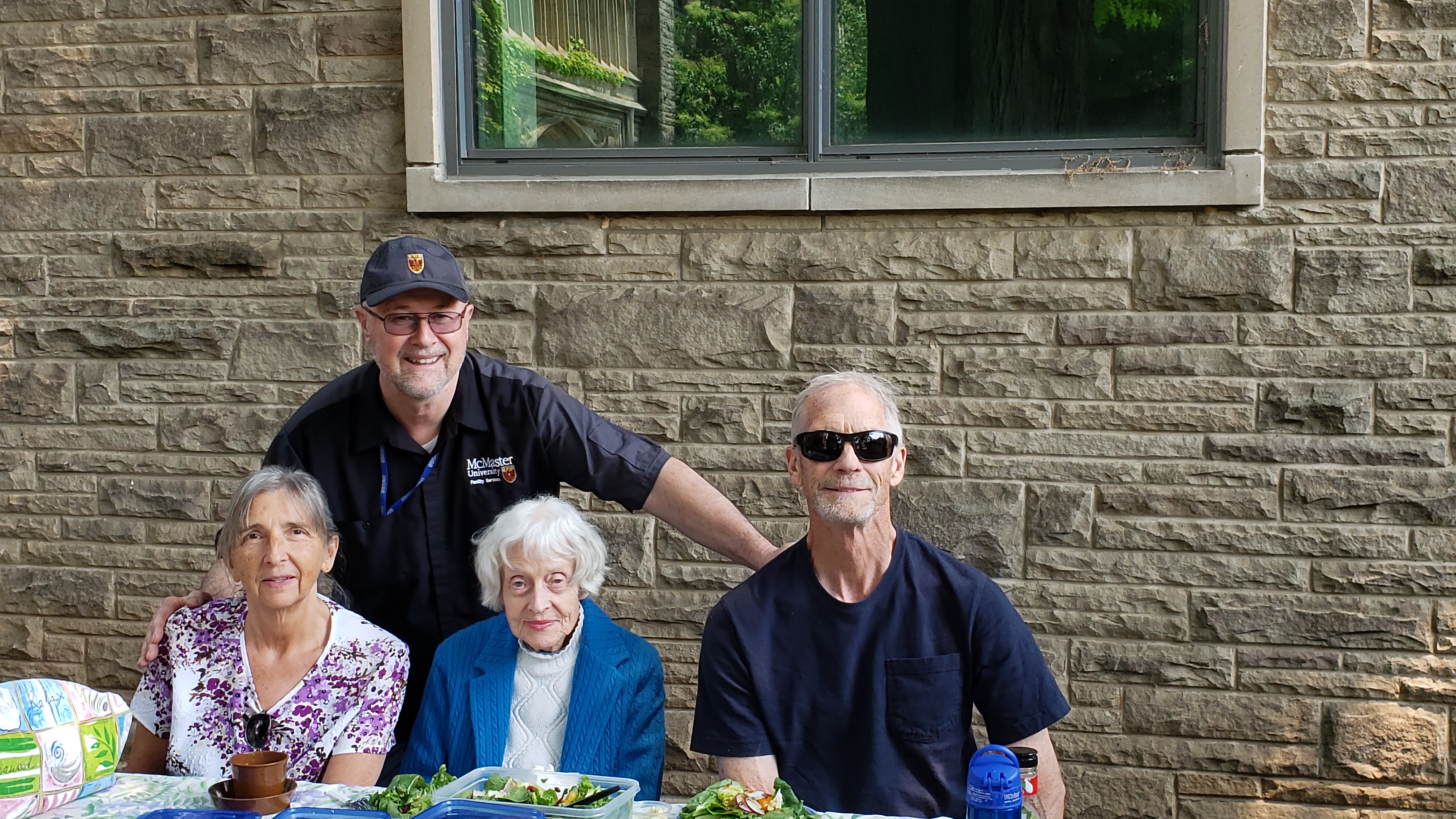 Three people sit at a lunch table, with a man standing behind them, in front of a stone building.