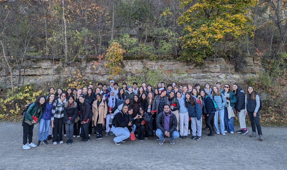 A large group of people stand outdoors, in front of a large rock and trees. 