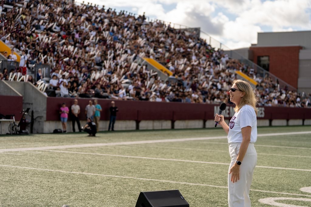 A laughing Maureen MacDonald holds the mic, standing on the football field, watched by thousands of cheering McMaster students in the stands at Mac Welcome.