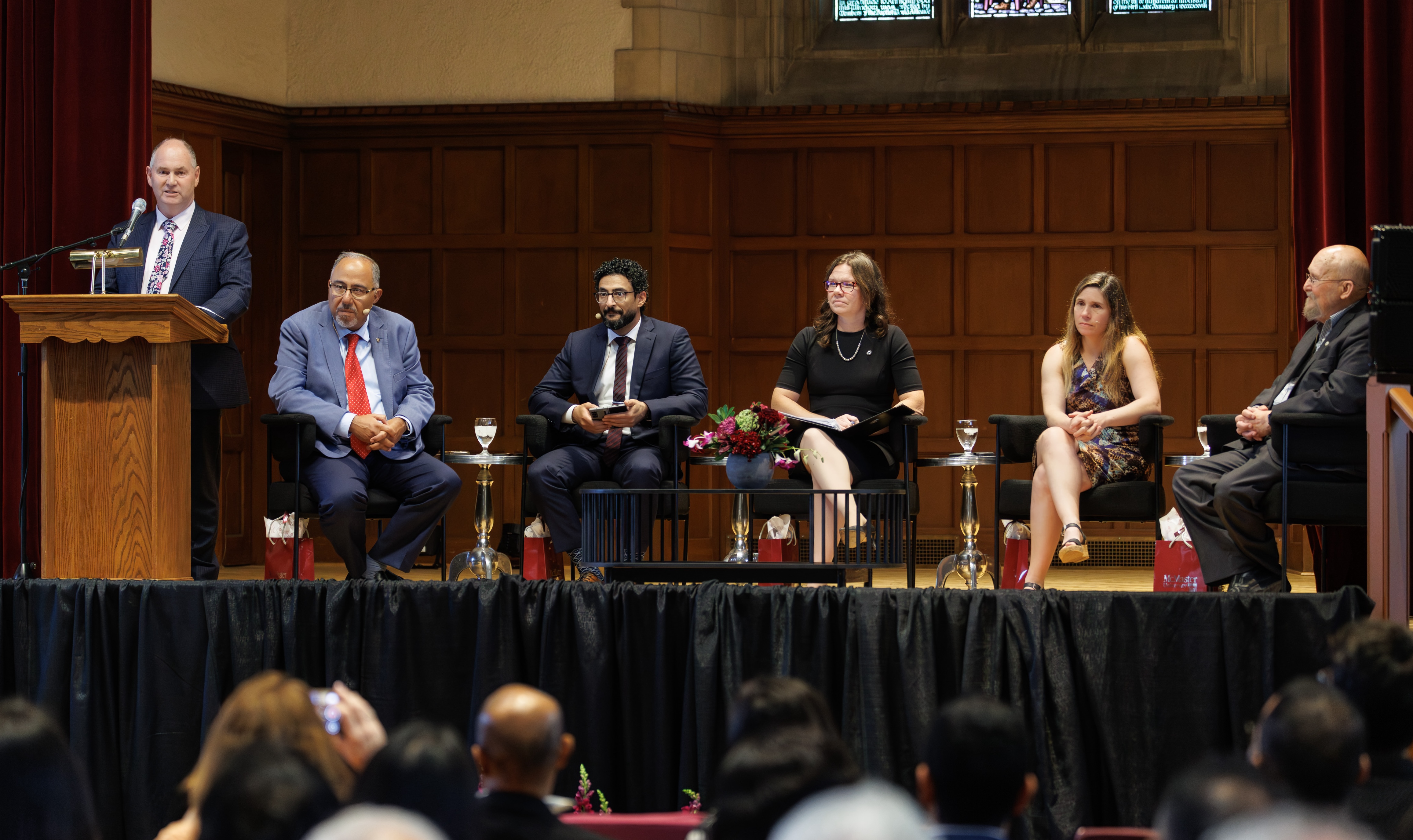A person in a suit speaks into a mic at the podium in Convocation Hall, watched by five panelists seated on stage.