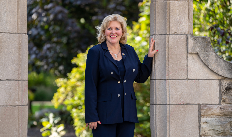 A woman in a navy suit places hand on archway at a university campus and looks to camera.