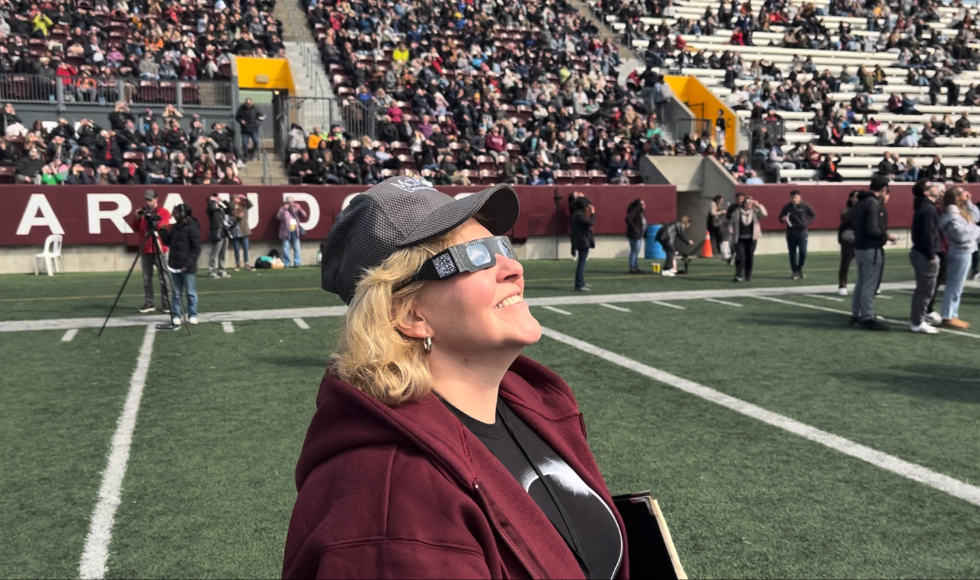 A person in a ball cap and maroon sweater, looks up to the sky, wearing solar eclipse glasses. A football stadium filled with spectators is in background.