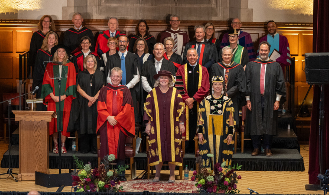 A smiling stage party dressed in academic regalia stands in four rows on the stage in Convocation Hall. President Susan Tighe is in the front row, centre.