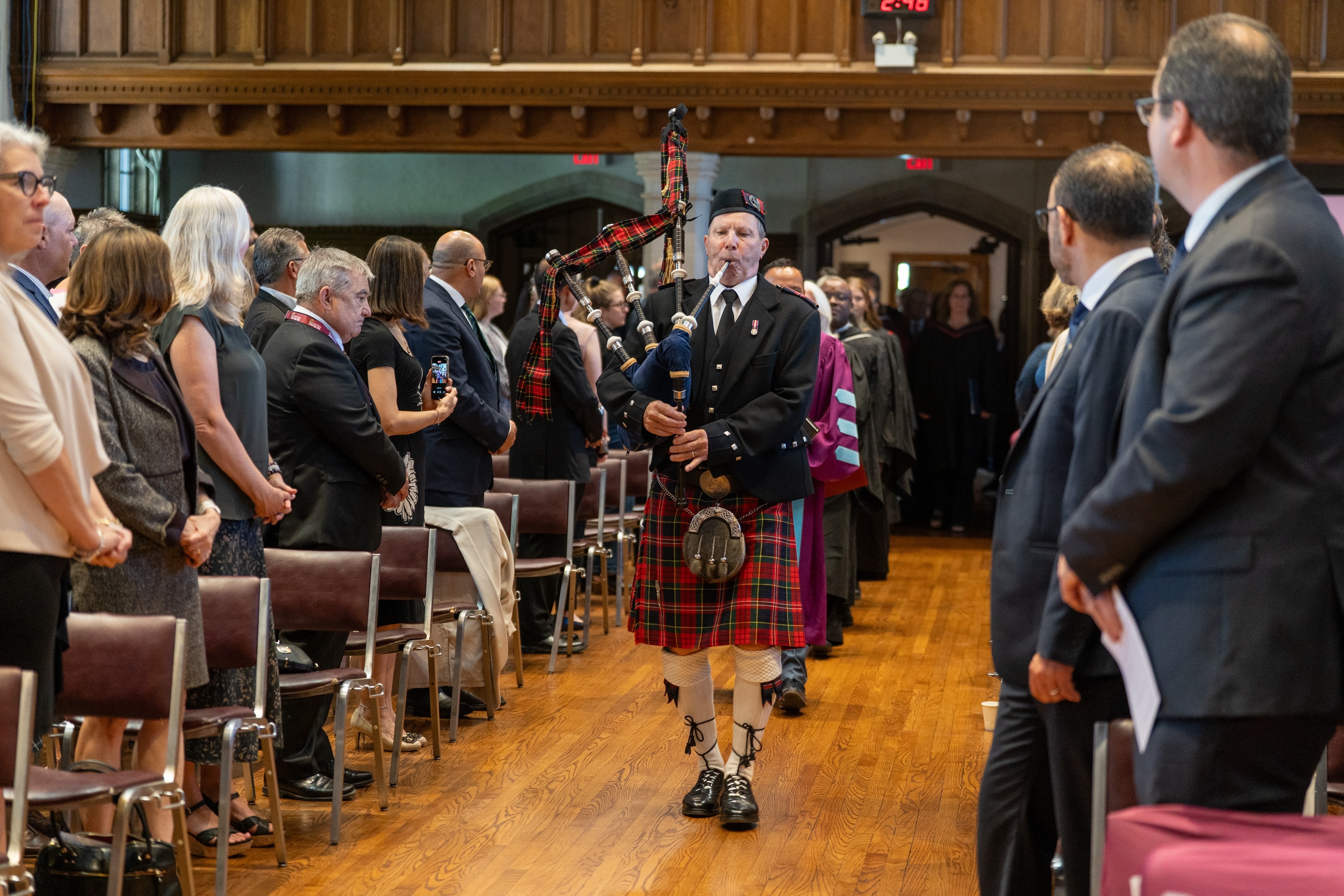 A man in a kilt plays the bagpipes as he leads a line of people down the centre of Convocation Hall, between two columns of attendees.