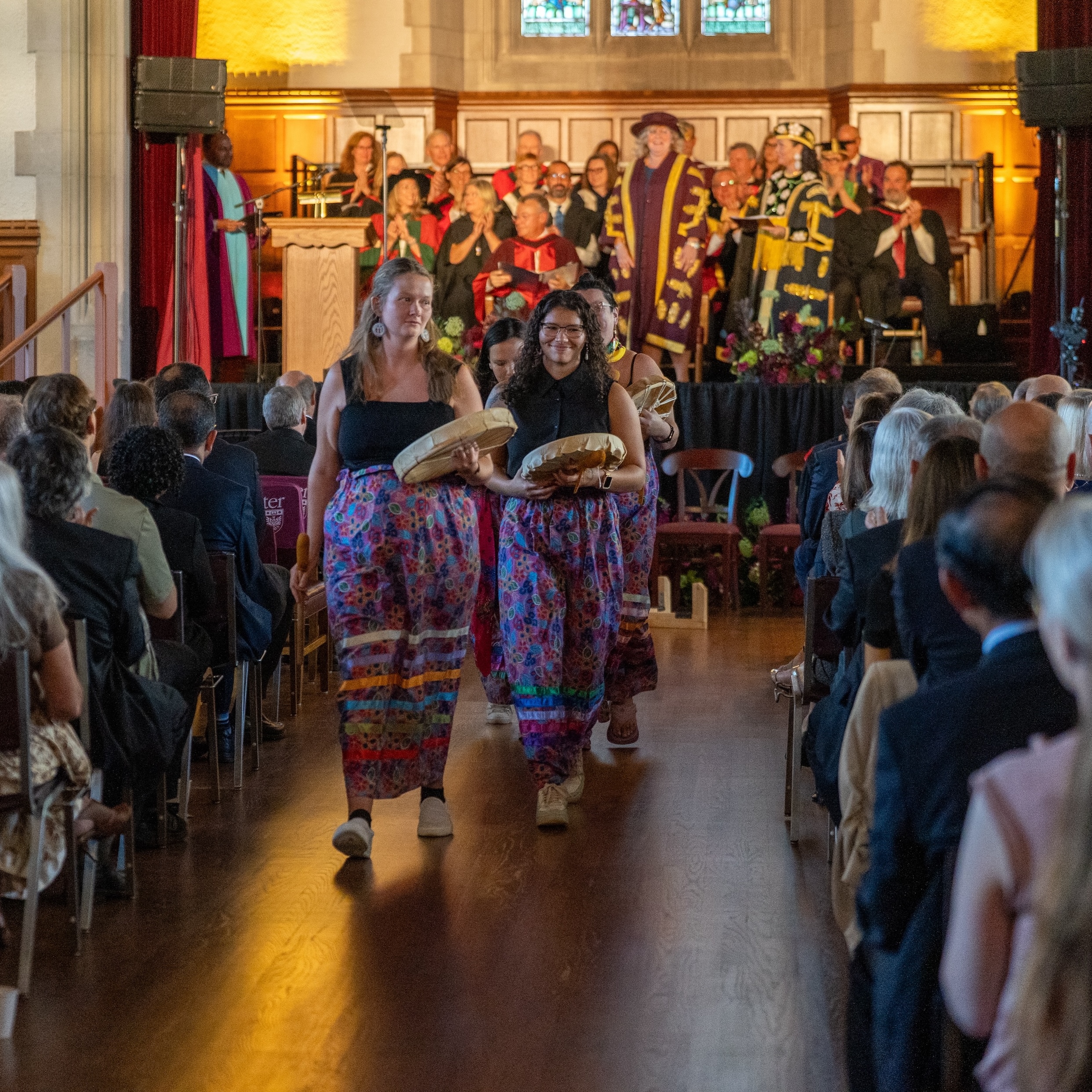 Women in ribbon skirts and holding traditional drums walk down the aisle in Convocation Hall, with the stage in the background.