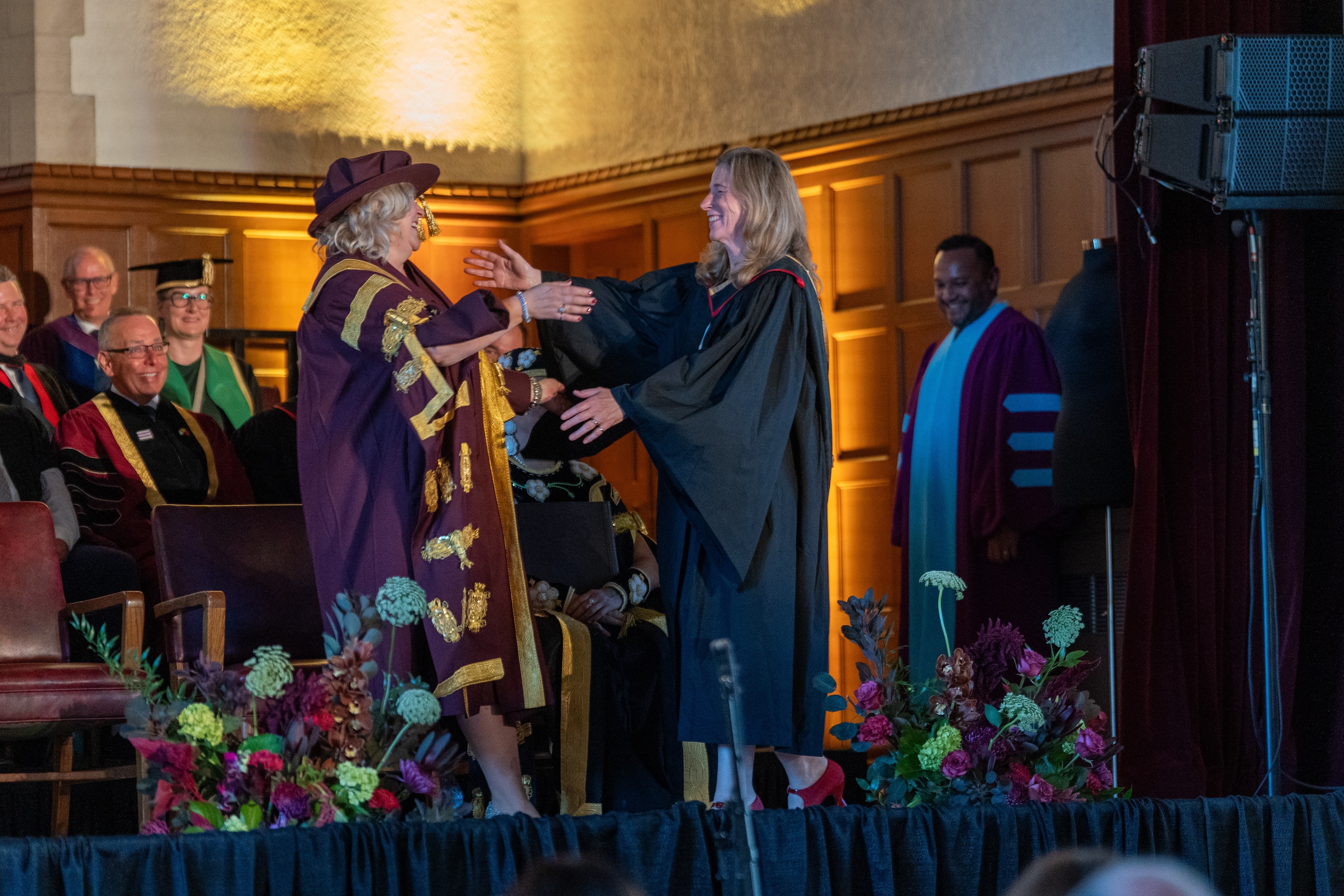 A professor in academic robes and President Susan Tighe raise their arms to hug at the installation ceremony.