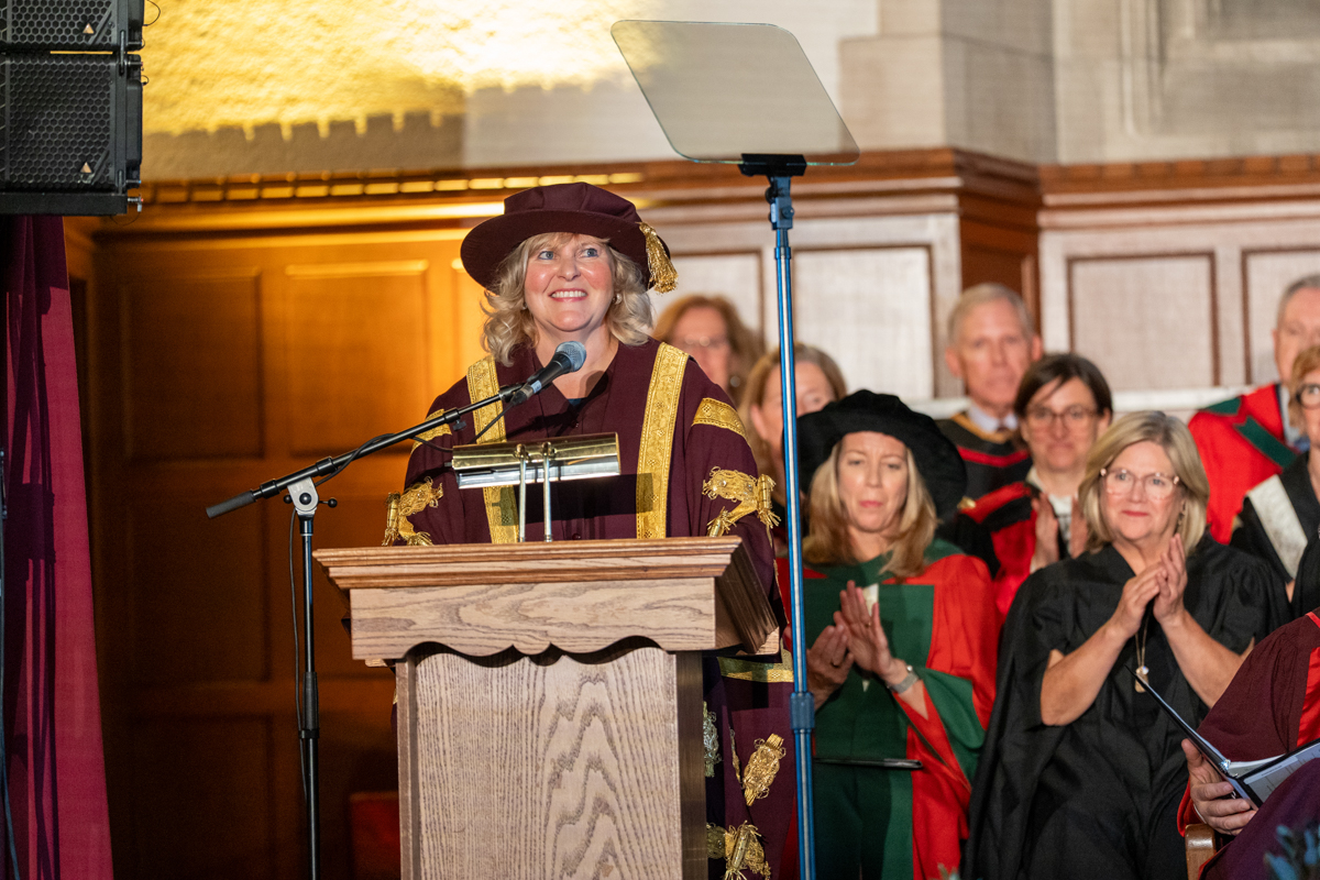 Susan Tighe, wearing the official presidential robes and hat, smiles as she speaks at the podium at her installation ceremony in Convocation Hall.