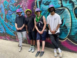 Four people wearing bucket hats in front of a spray painted brick wall