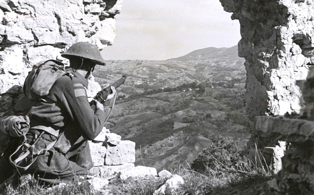 hole blasted through a hill-top wall in Italy gives this Canadian a vantage point from which to observe enemy movements while men of his unit move into a new position.