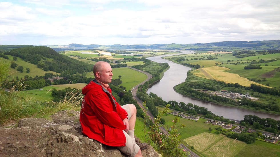 Jamie Tennant, in a red jacket, sits on a hill in Scotland