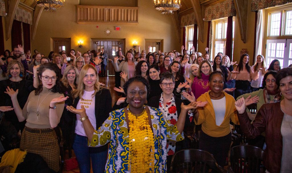 A room packed with women and a few men strike a pose with their hands up to represent 