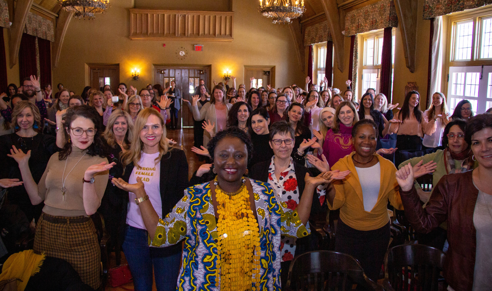 A room packed with women and a few men strike a pose with their hands up to represent