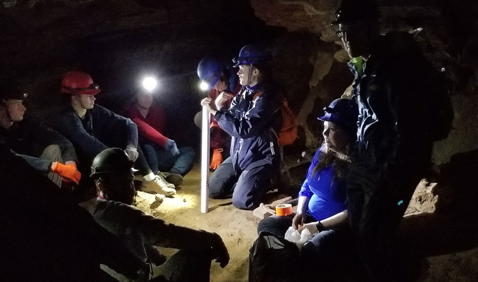 Students wearing caving equipment sit in a circle, lit by a lantern.
