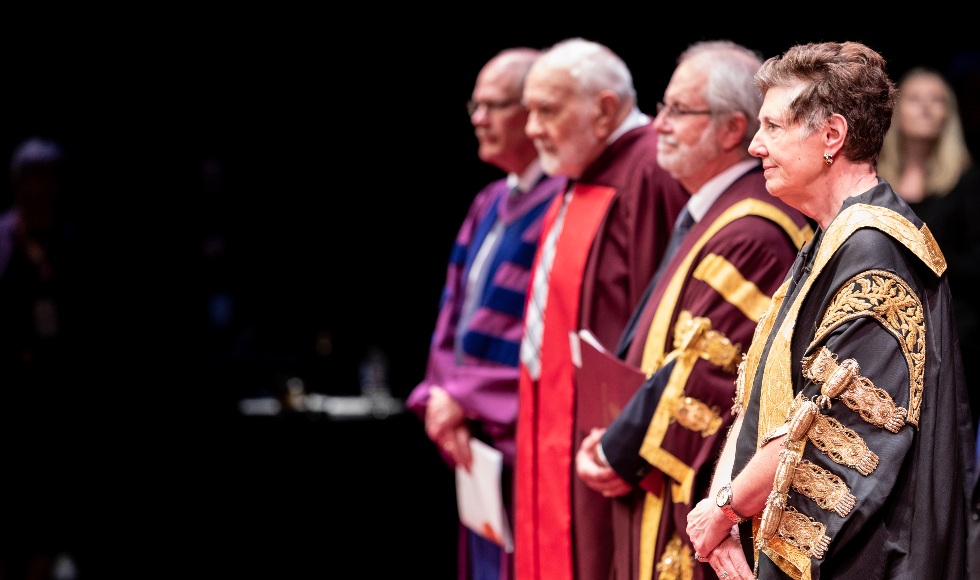 Three men and one woman stand in academic regalia onstage at McMaster's HUmanities convocation in 2019.