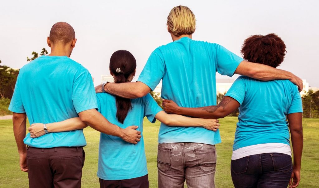 Photo from behind of four people with their arms around each other's waists.