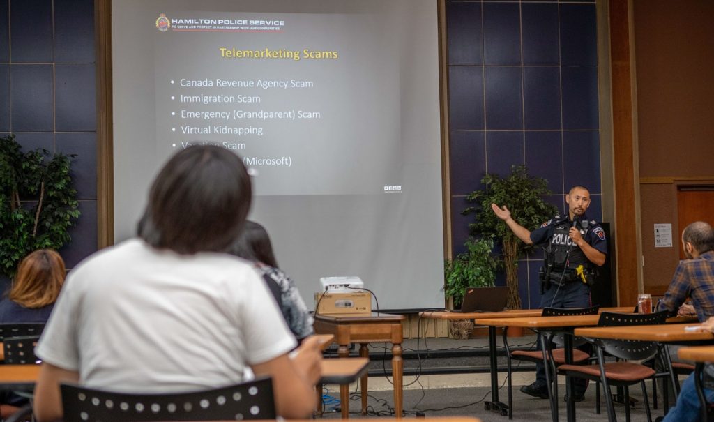 Police officer stands in front of a large slide listing examples of telemarketing scams.