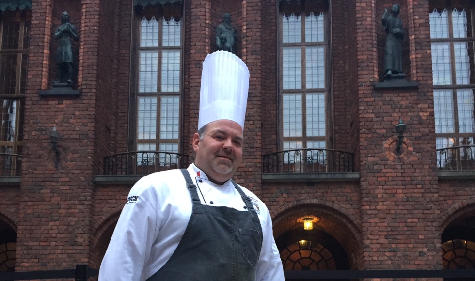 A man wearing a tall chef's hat and a black apron stands in front of Stockholm City Hall