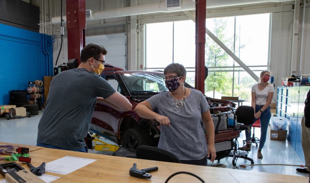 Two people wearing masks bump elbows in a workshop. Behind them is a car with its hood popped open, lots of machinery and tools everywhere.