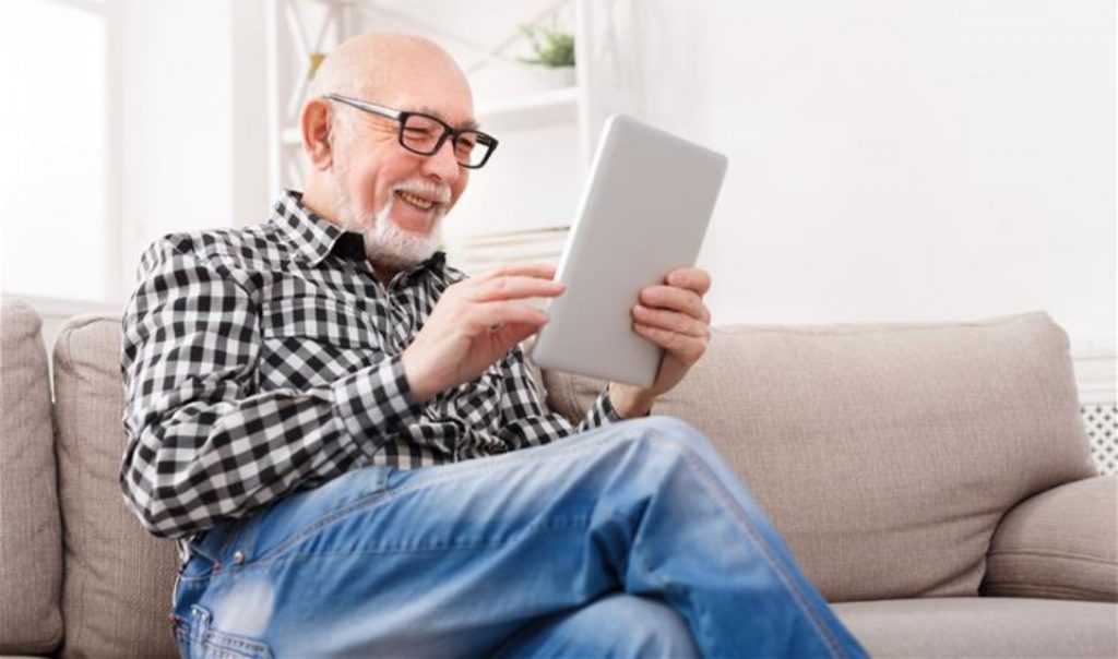 An older man with a short white beard sitting on a couch and smiling at a tablet.