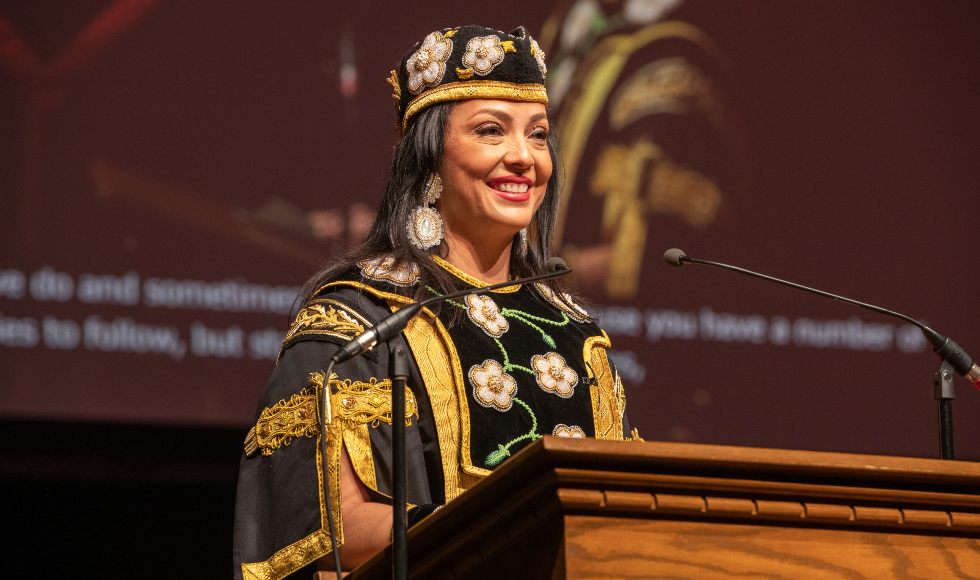 A woman in beaded robes stands at a lectern