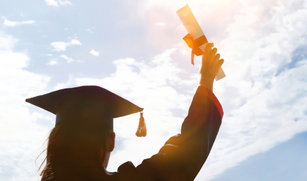 A young woman, seen from the back, in a graduation cap and gown holding a diploma