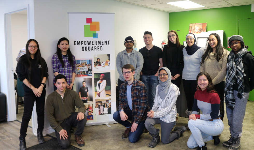 A multi-racial group of students stands around an Empowerment Squared banner in a room with a birght green wall