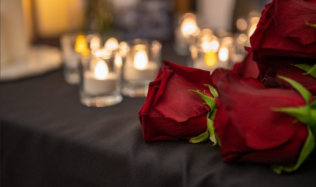 Roses lie on a table in front of several lit votive candles. This image is from a memorial vigil held in January 2020 for those killed in the Tehran plane crash.