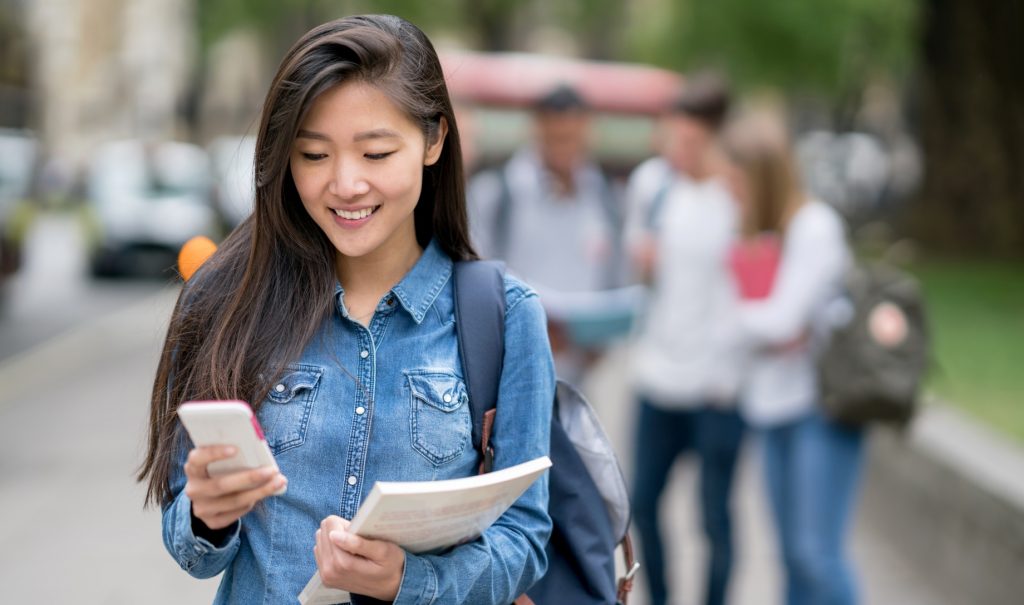 A woman with a backpack and papers smiles down at her phone
