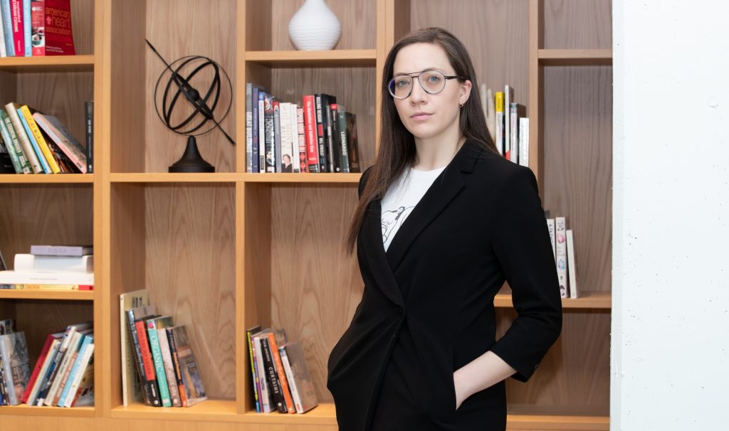 Vass standing in front of a bookcase with her hands in her blazer pockets.