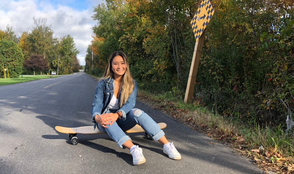 Kaitin Jingco sitting on a skateboard on an open road, smiling at the camera