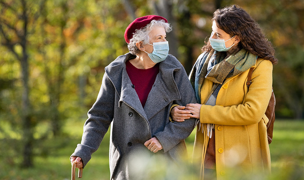 An older adult with white hair and a red hat using a cane is walking with a younger woman in a yellow coat who is holding her arm