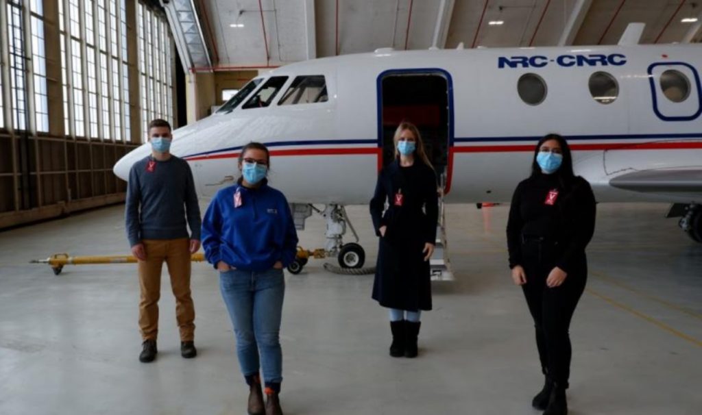 Four students wearing masks and standing distanced in front of an airplane in a hangar.