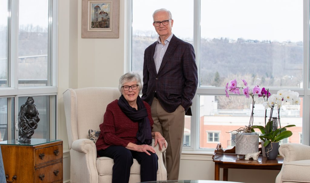 A smiling woman sitting in an armchair in a well-lit room, with a smiling man standing beside her.