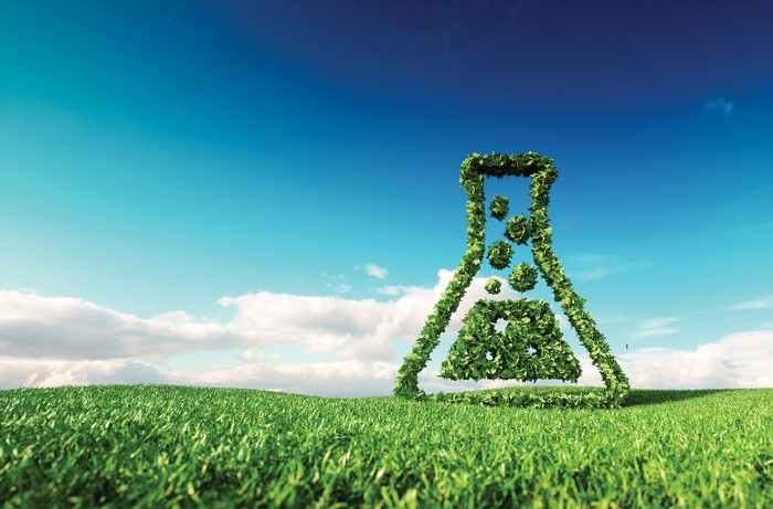 Greenery in the shape of a beaker set against a backdrop of a blue sky and a field of green grass.