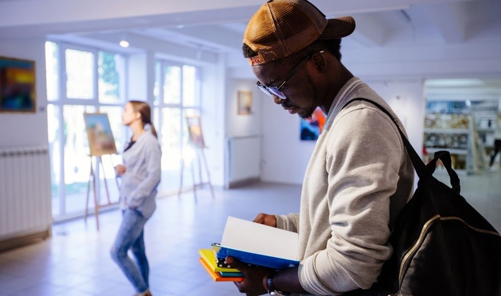 Young male in a sweatshirt and ball cap looking down at the books in his hands while standing in a room full of art and easels.