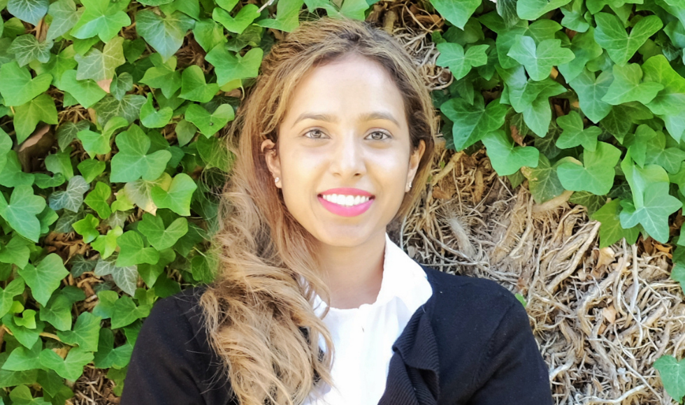 A photo of Allison Leanage smiling at the camera. She is photographed from the chest up and wearing a white shirt and black cardigan. She is positioned directly in front of some vines and greenery.