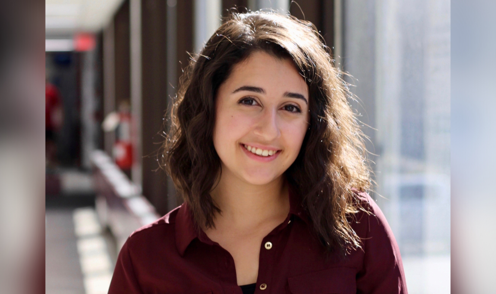 A photo of Claudia Turco from the shoulders up. She is wearing a maroon button-up shirt and smiling at the camera. The background is out of focus and she is standing in a hallway with alot of natural light.