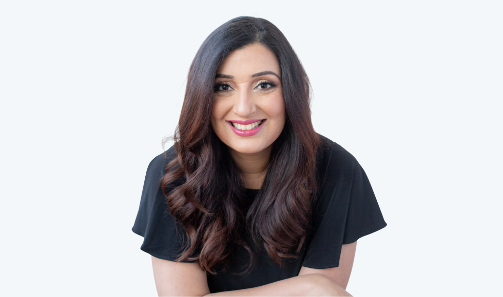 A picture of Samra Zafar from the waist up, smiling at the camera. She is wearing a black, short-sleeved shirt and is against a white backdrop.